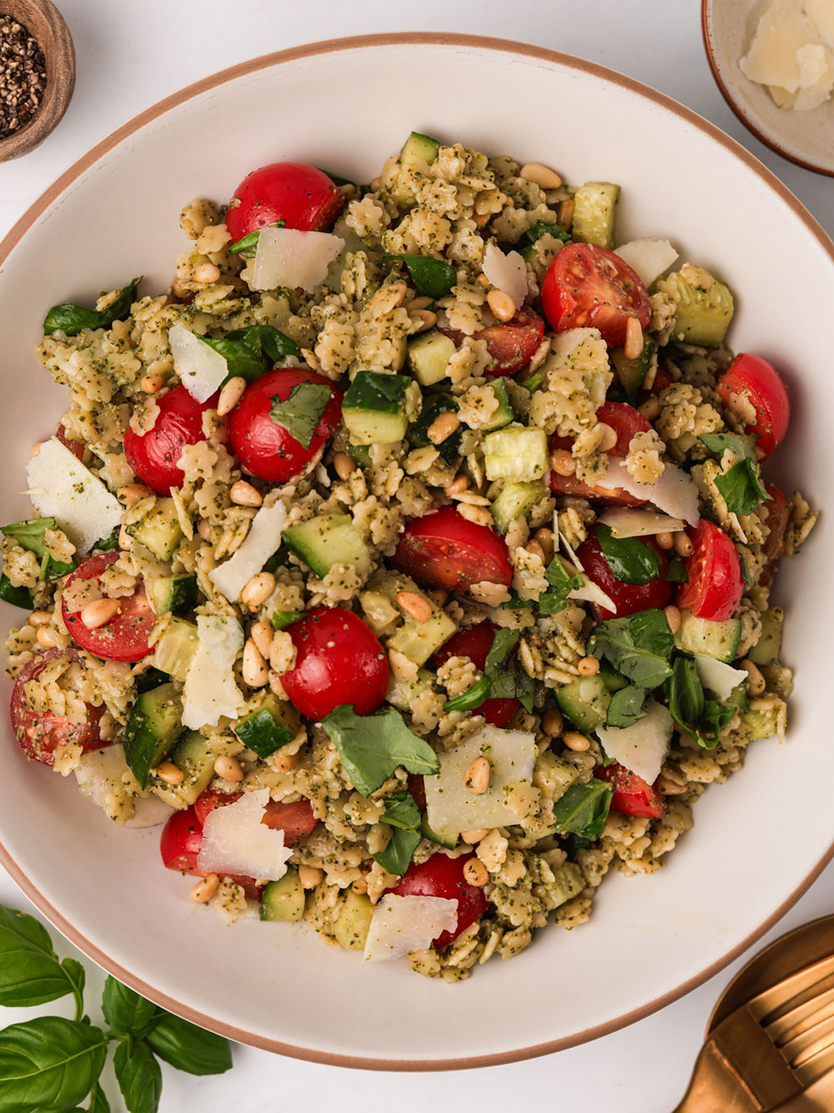 A serving of Pastina Salad in a white brown rimmed bowl with basil, parmesan and black pepper on the side.
