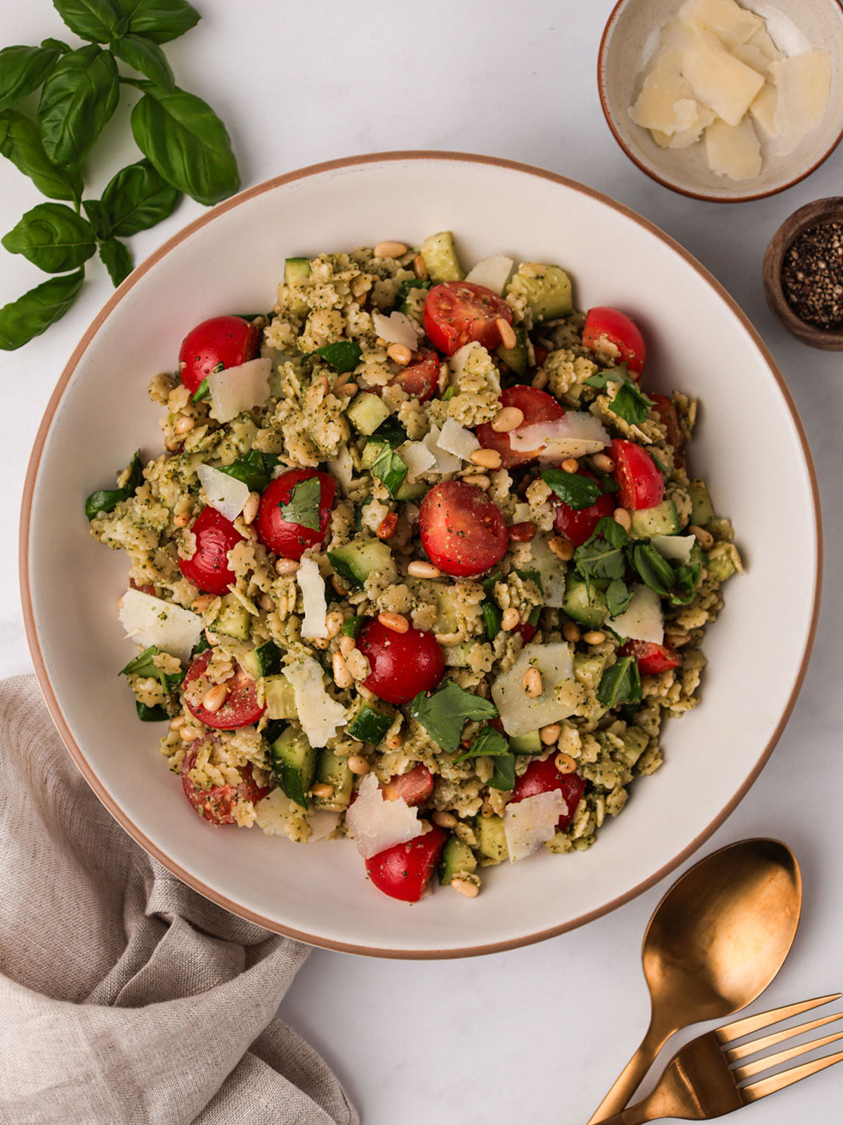 A serving of Pastina Salad in a white brown rimmed bowl with basil, parmesan and black pepper on the side.