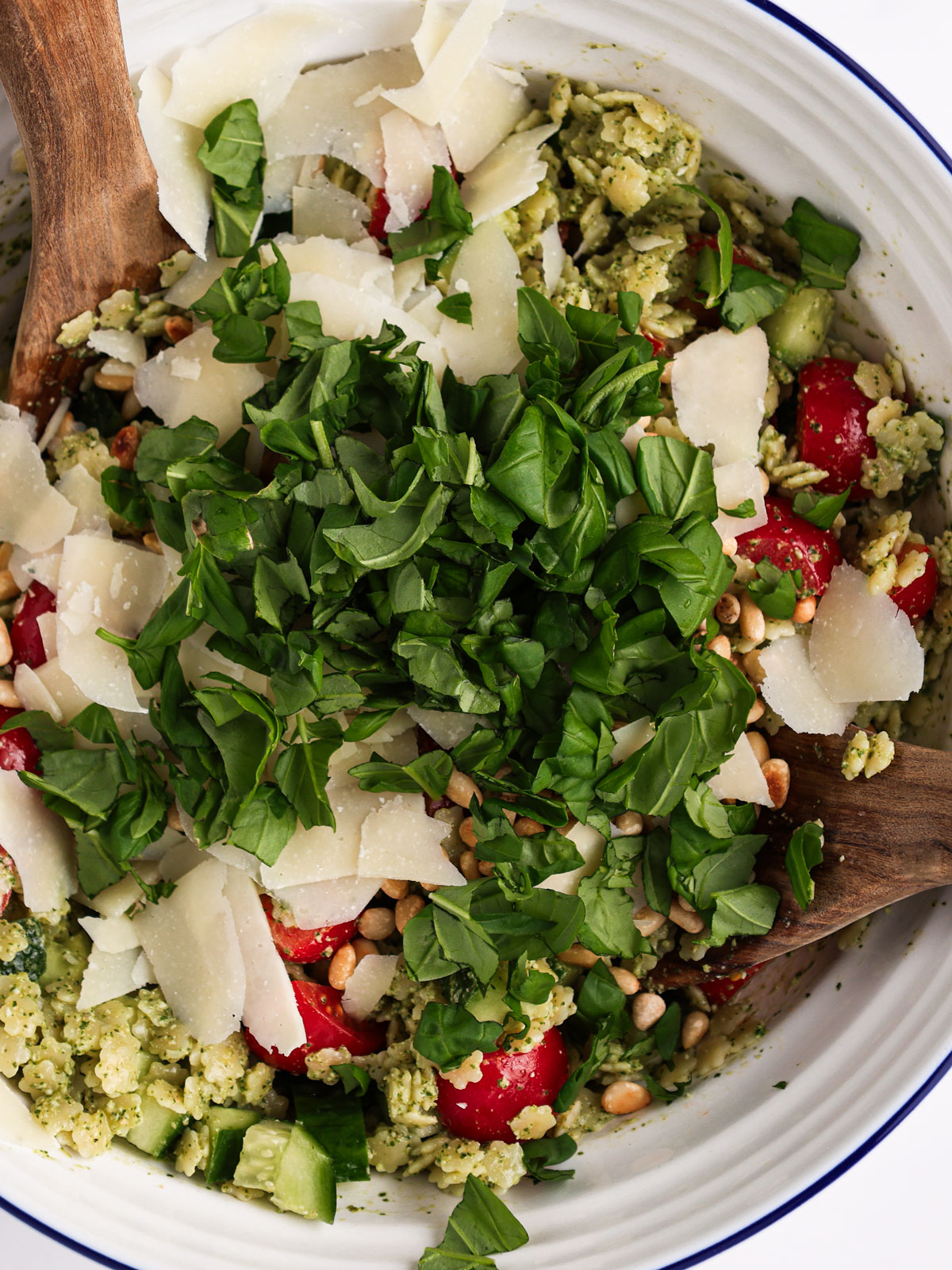 Folding in the tomatoes, cucumber, and basil, then topping with Parmesan shavings and toasted pine nuts. In a white blue rimmed bowl.